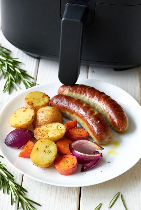 Golden brown sausages with roasted carrots, baby potatoes, and red onion on a white ceramic plate, air fried to perfection